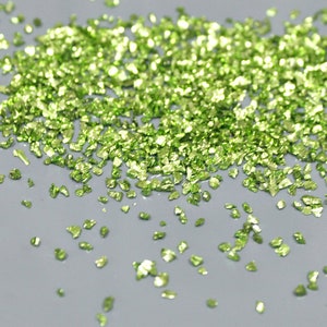 May include: A scattering of small, irregularly shaped, light green glass pieces. The fragments are scattered across a gray surface, catching the light and reflecting a bright, shimmering appearance. The overall effect is of a sparkling, textured material.
