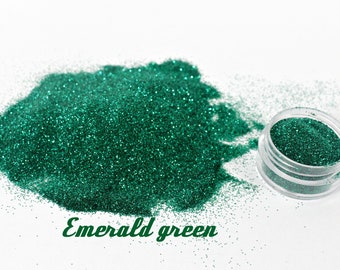 Puede incluir: Purpurina verde esmeralda esparcida sobre una superficie blanca, con un pequeño recipiente transparente lleno de la misma purpurina. El texto "Emerald green" está escrito en cursiva debajo.