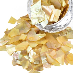 May include: A pile of iridescent, light yellow, and gold-toned, irregular shaped, mother of pearl shell pieces. The shell pieces are scattered on a white background.