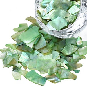 May include: A bowl of iridescent green and gold shell pieces. The shell pieces are various sizes and shapes, and they are scattered on a white surface.