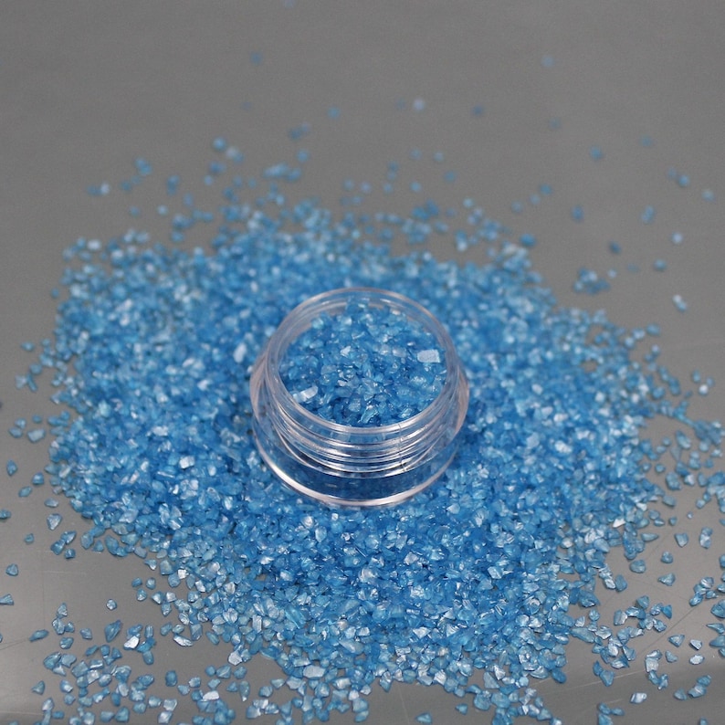 May include: A small, clear, circular plastic container filled with tiny, bright blue, angular, crystalline fragments. A large pile of the same blue, crushed, glass-like particles is scattered around the base of the open jar.
