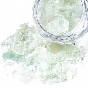 Może przedstawiać: Szklana miska wypełniona zielonymi płatkami miki, z dodatkowymi płatkami rozrzuconymi wokół miski na białym tle.