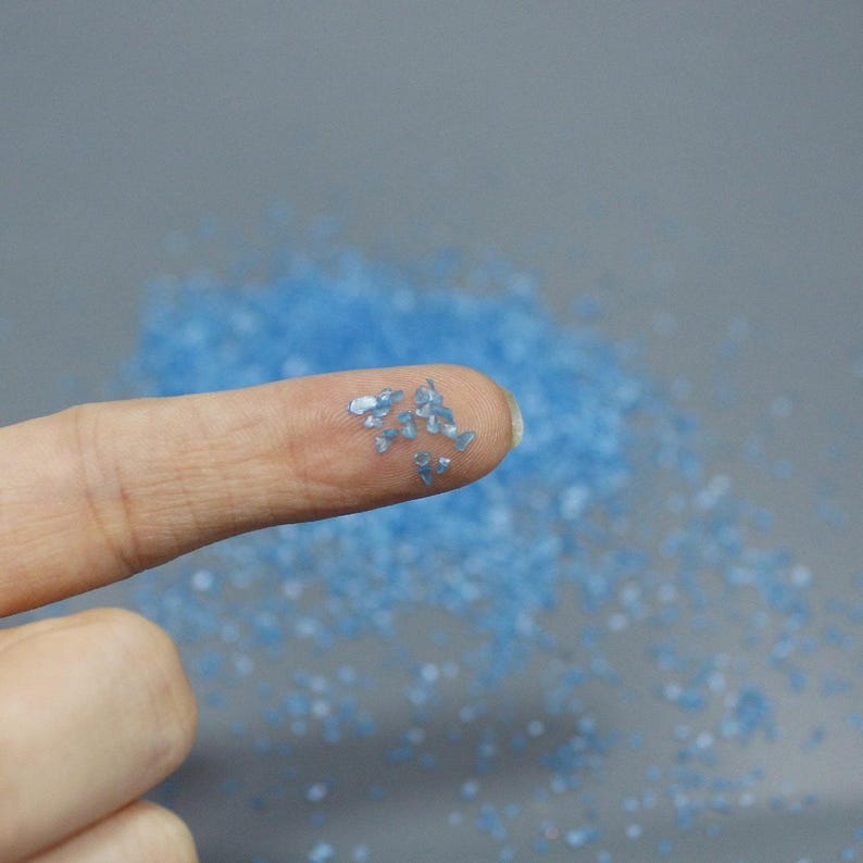 May include: A collection of small, light blue, irregular, translucent, and reflective glass or mineral fragments. The tiny, jagged pieces are scattered across a surface, with a few individual shards resting on a fingertip.