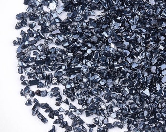 May include: A close-up view of a pile of small, dark gray, iridescent glass chips. The chips are scattered on a white surface.