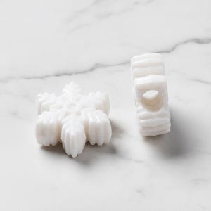 May include: Two white snowflake-shaped beads with a glossy finish. One bead is lying flat, displaying its six-pointed star design, while the other is standing on its side, revealing a central hole running through the middle of the bead.