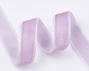May include: A length of lavender velvet ribbon, with a soft, plush texture. The ribbon is folded in a gentle curve, showcasing its delicate color and sheen. The ribbon's edges are a lighter shade of lavender.