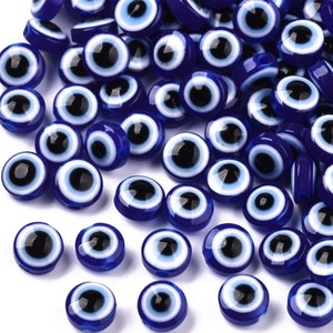 May include: A collection of blue and white evil eye beads. The beads are round and have a white center with a black pupil. The beads are scattered on a white surface.