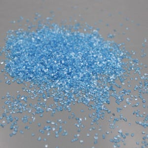 May include: A pile of small, light blue crystalline granules with a reflective, faceted texture. The individual particles are irregular in shape and size, scattered across a smooth, neutral-toned surface, creating a granular appearance.
