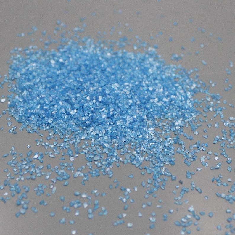 May include: A pile of small, light blue crystalline granules with a reflective, faceted texture. The individual particles are irregular in shape and size, scattered across a smooth, neutral-toned surface, creating a granular appearance.