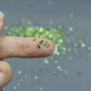 May include: Close-up of a finger holding small, shiny, green glitter flakes. The flakes are scattered on the finger and in the background, creating a sparkling effect. The glitter appears to be metallic.
