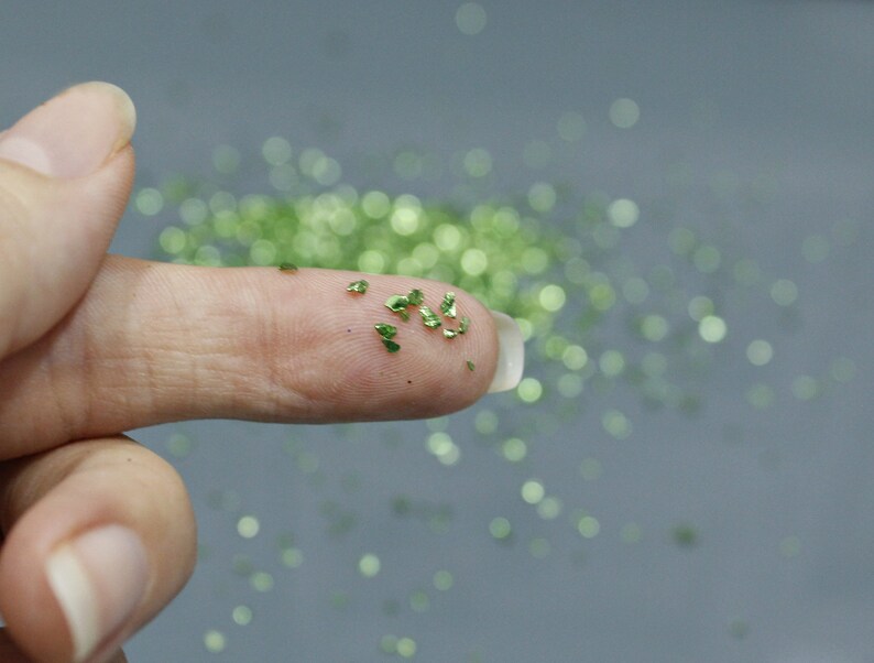 May include: Close-up of a finger holding small, shiny, green glitter flakes. The flakes are scattered on the finger and in the background, creating a sparkling effect. The glitter appears to be metallic.