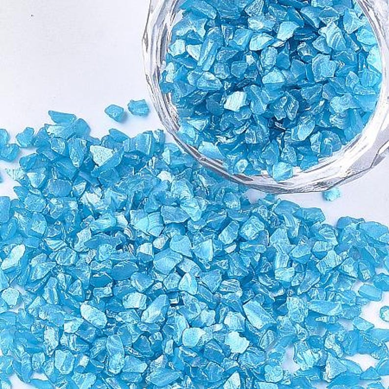 May include: A collection of small, irregular, bright blue glass shards or crushed stones. Some of the blue fragments are contained within a clear glass bowl, while others are scattered across the surface in a loose, textured pile.