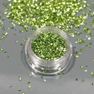 May include: A small, clear plastic jar filled with bright green, crushed glitter. The glitter is scattered around the jar on a gray surface. The glitter has a reflective, metallic appearance.