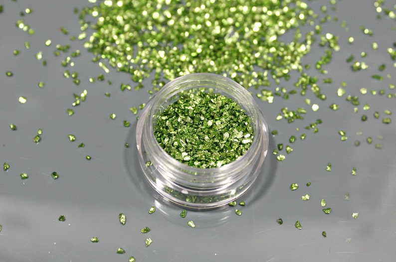 May include: A small, clear plastic jar filled with bright green, crushed glitter. The glitter is scattered around the jar on a gray surface. The glitter has a reflective, metallic appearance.