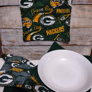 May include: A square quilted pot holder and a bowl cozy featuring a dark green fabric with a repeating Green Bay Packers logo, football helmet, and text pattern. A plain white ceramic bowl sits inside the fabric bowl cozy, which has a matching green interior.