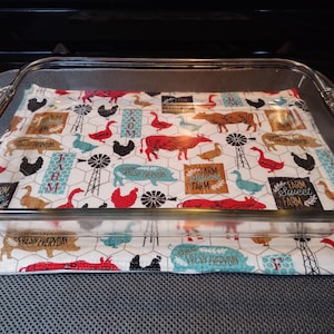 May include: A clear glass baking dish with a white fabric liner. The liner has a farmhouse theme with a repeating pattern of chickens, pigs, cows, windmills, and the text "Farm Sweet Farm".