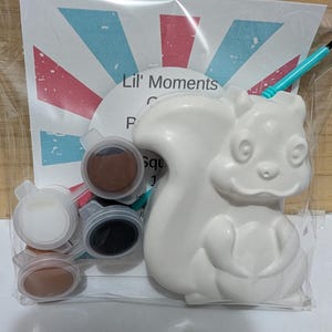May include: A craft kit in a clear plastic bag containing a white ceramic squirrel figurine, a small paintbrush with a teal handle, and four small circular paint pots in white, black, and two shades of brown, with a card labeled Lil' Moments.
