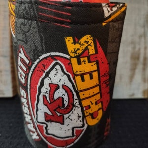 May include: A black and red can cooler with a distressed Kansas City Chiefs logo. The cooler has a yellow "Chiefs" text and a red and white arrowhead logo. The cooler is made of fabric and has a stitched seam around the top.