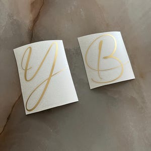 Könnte beinhalten: Zwei weiße Aufkleber mit goldenen Schreibschriftbuchstaben, ein "Y" und ein "B". Die Aufkleber befinden sich auf einer neutral getönten Oberfläche. Die goldene Schrift hat ein glänzendes Finish.