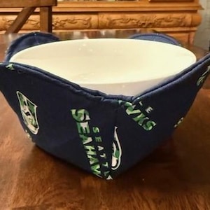May include: A blue and green bowl cozy with the Seattle Seahawks logo. The cozy is made of fabric and has a white bowl inside.