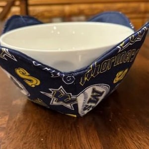 May include: A blue and yellow fabric bowl cozy with a baseball and the word "Mariners" printed on it. The cozy is holding a white ceramic bowl.