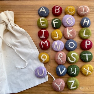 May include: A set of 26 felt alphabet letter stones in various colors, including red, green, blue, yellow, purple, and brown. The stones are arranged in alphabetical order and are contained in a white drawstring bag.