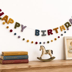 May include: A felt Happy Birthday banner featuring multicolored letters and small felt balls strung on a white cord. The letters are in shades of red, yellow, olive green, purple, and dark blue, creating a festive garland for celebrations.
