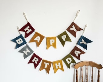 May include: A colorful felt banner spelling "HAPPY BIRTHDAY" in white letters. The pennants are in various colors, including red, blue, yellow, and brown, strung together with white cord. A wooden chair is visible in the bottom right corner.
