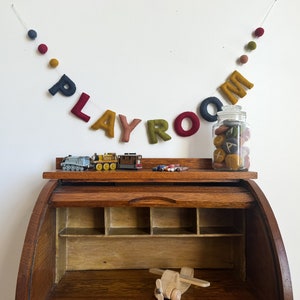 May include: A felt letter banner spelling "PLAYROOM" hangs above a wooden desk with toy trains and a wooden airplane. The banner is made of felt letters in various colors, including red, yellow, green, and blue. The letters are strung on a white string with felt balls in between.