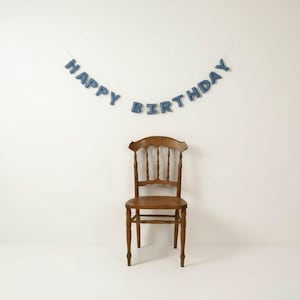 May include: A wooden chair with a curved back and a woven seat is positioned in front of a white wall. Above the chair, a banner spells out "HAPPY BIRTHDAY" in blue letters, creating a festive atmosphere. The chair has a classic design.