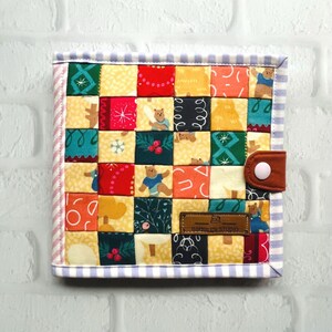 May include: A patchwork fabric notebook cover with a variety of colorful patterns, including bears, trees, and snowflakes. The cover has a brown leather strap with a white button closure and a brown leather label that reads "Dappled Studio".