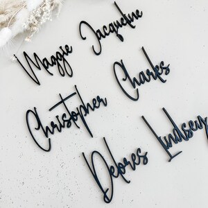 K&ouml;nnte beinhalten: Schwarze Acryl-Platzkarte mit den Namen Maggie Jacqueline, Christopher Charles, Delores Lindsey in kursivschrift.