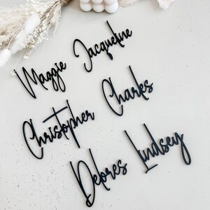 K&ouml;nnte beinhalten: F&uuml;nf schwarze Acryl-Namen, "Maggie Jacqueline", "Christopher Charles", "Debores Lindsey", werden auf einer hellgrauen Oberfl&auml;che angezeigt. Die Namen sind in kursiver Schrift geschrieben.