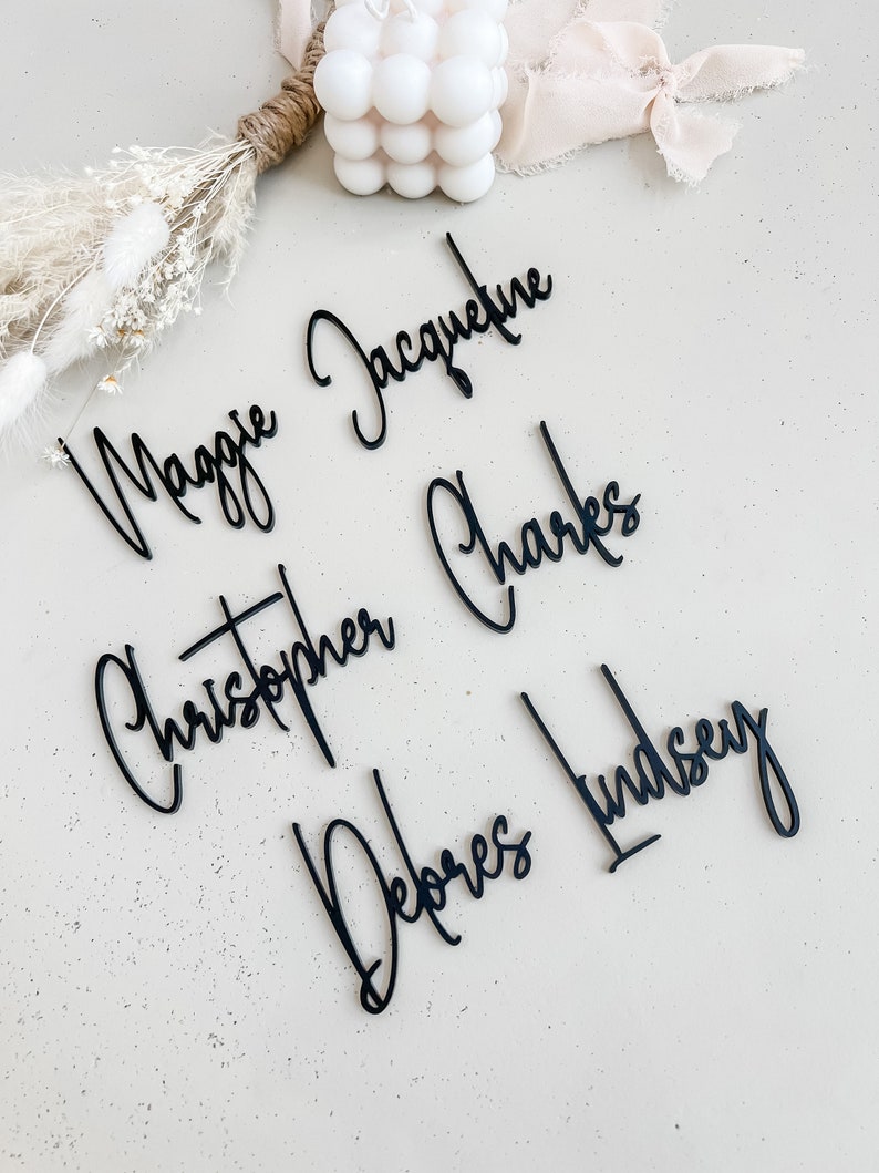 K&ouml;nnte beinhalten: F&uuml;nf schwarze Acryl-Namen, "Maggie Jacqueline", "Christopher Charles", "Debores Lindsey", werden auf einer hellgrauen Oberfl&auml;che angezeigt. Die Namen sind in kursiver Schrift geschrieben.