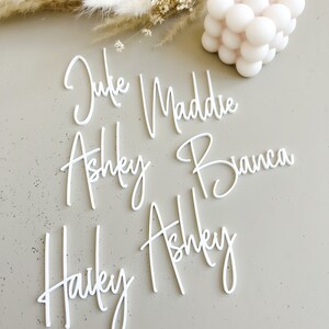 K&ouml;nnte beinhalten: Wei&szlig;e Acryl-Namen, darunter "Julie", "Maddie", "Ashley", "Bianca" und "Hailey", sind auf einer hellgrauen Oberfl&auml;che angeordnet.