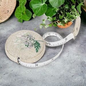 May include: A white fabric measuring tape with a floral design on the case. The tape is rolled up and the case is made of hessian.