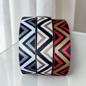 May include: Three patterned straps with a zig-zag design. The straps feature black, white, grey, and tan colours. The straps are likely for a bag or other accessory.