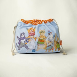 May include: A light blue drawstring bag featuring a colorful illustration of several cats wearing winter clothing, including scarves, hats, and sweaters. The top edge has an orange fabric trim with a white pattern. An orange tag reads Emma Ball.
