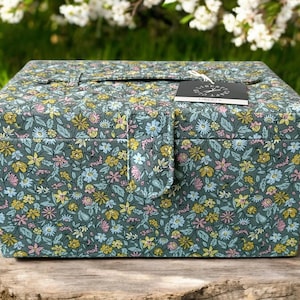 Op de afbeelding: Een vierkante doos met bloemenpatroon, een scharnierend deksel en een sluiting. De doos is bedekt met een stof met een donkergroene achtergrond en een patroon van kleine gele, roze en blauwe bloemen. Een label is bevestigd.