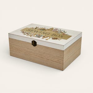 May include: A rectangular wooden sewing box with a light brown base and a white lid. The lid features the words "SEWING BOX" in gold lettering, along with illustrations of sewing tools. A small, dark metal clasp secures the box.