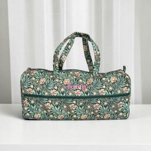 Può includere: Una borsa da viaggio verde a fantasia floreale con chiusura a zip e manici. La borsa presenta un motivo di fiori e foglie nei toni del verde, rosa e crema. Il nome "Jenny" è ricamato in rosa sul davanti.