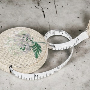 May include: A white fabric measuring tape with a floral design. The tape is rolled up and the end is attached to a small, round, fabric case with a floral design.