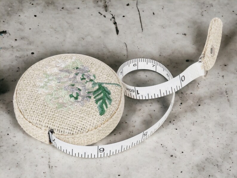 May include: A white fabric measuring tape with a floral design. The tape is rolled up and the end is attached to a small, round, fabric case with a floral design.