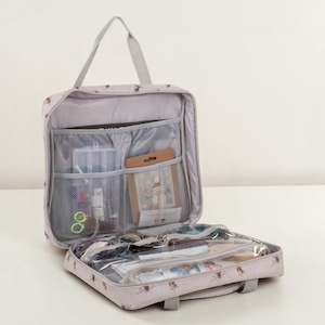 May include: Open, pale grey travel organiser with a handle and multiple compartments. The organiser is filled with various craft supplies, including small containers, spools of thread, and other sewing notions. The exterior features a subtle pattern of small brown floral designs.