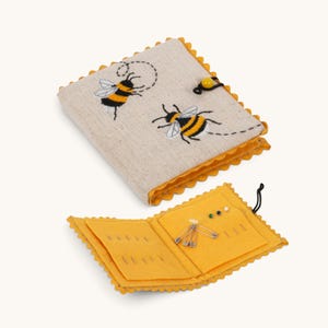 May include: A square needle book with a beige linen cover featuring two embroidered bumblebees and a yellow bead closure. The interior is made of yellow felt with scalloped edges, containing several metal safety pins and sewing needles.
