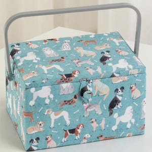 May include: A rectangular sewing basket with a teal fabric exterior featuring a repeating pattern of various dog breeds and small white bones. The basket includes a gray plastic handle and a fabric-covered button closure on the front flap.