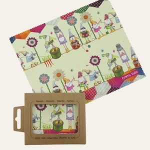 May include: A microfiber lens cloth with a whimsical gnome pattern in a box. The cloth features gnomes, flowers, and other garden elements on a light green background. The box is brown with a clear window showing the cloth design. The text "Emma Balle" is visible.