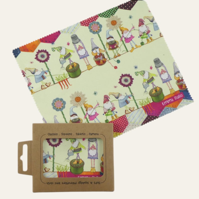 May include: A microfiber lens cloth with a whimsical gnome pattern in a box. The cloth features gnomes, flowers, and other garden elements on a light green background. The box is brown with a clear window showing the cloth design. The text "Emma Balle" is visible.
