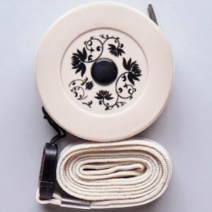 May include: Cream-coloured tape measure with a black floral design on the circular casing. The tape measure has a black button in the centre and a rolled-up beige measuring tape. A black strap is attached to the casing.
