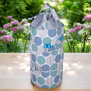 May include: A gray and white cylindrical knitting bag with a drawstring top and a handle. The bag has a pattern of yarn balls and is filled with knitting needles.